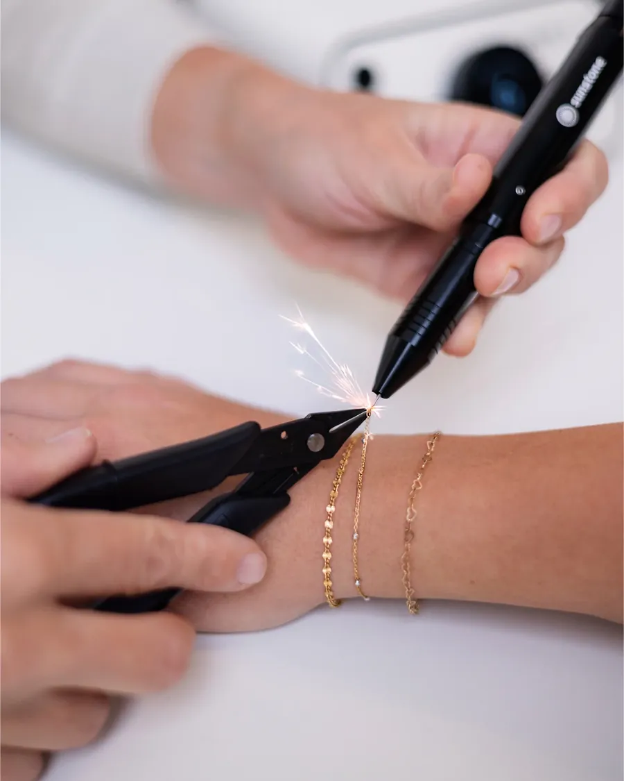 Primer plano de una pulsera de cadena fina de oro soldándose en la muñeca de una clienta, con una pequeña chispa; la artista sostiene el estilete y unas pinzas.